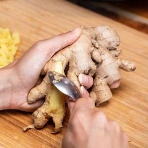Frische Ingwerwurzel wird mit einem Löffel geschält, daneben frisch geriebener Ingwer